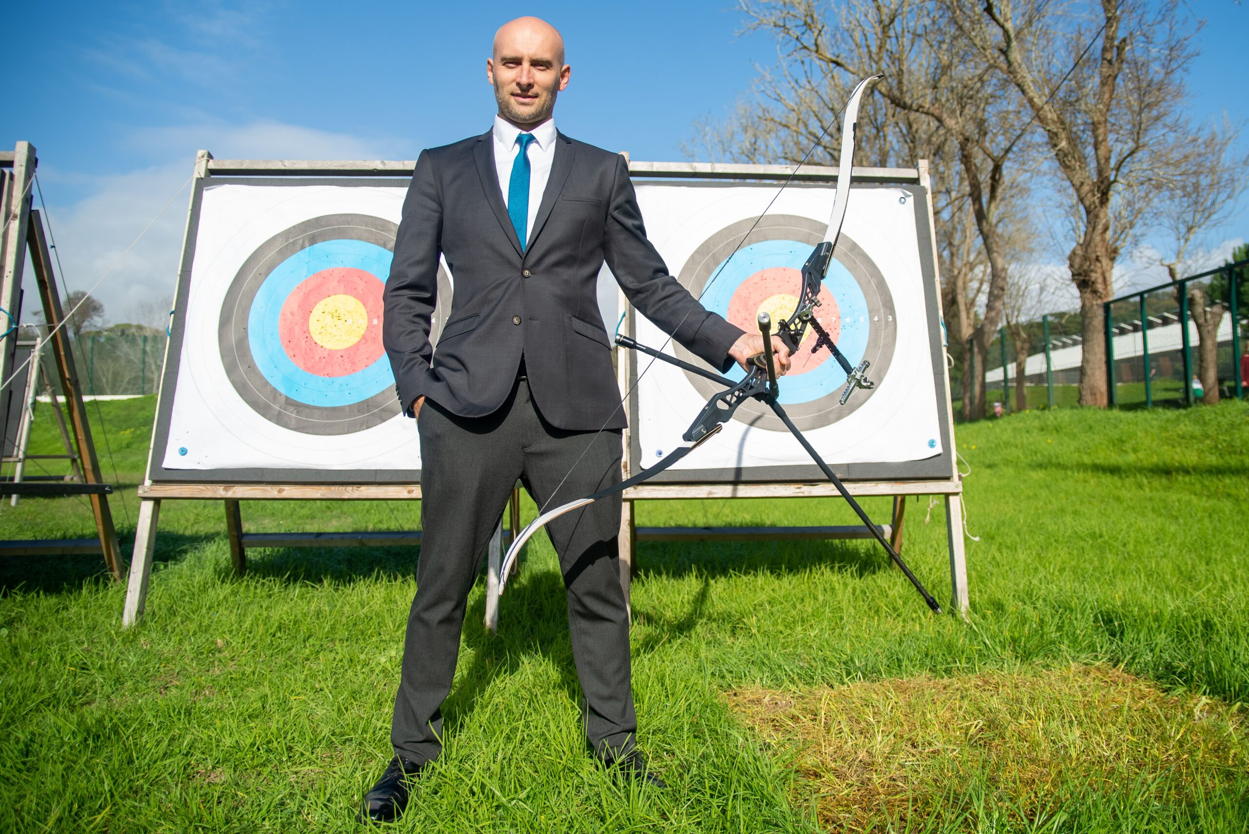 A business professional in a suit practicing archery with targets outdoors on a sunny day.